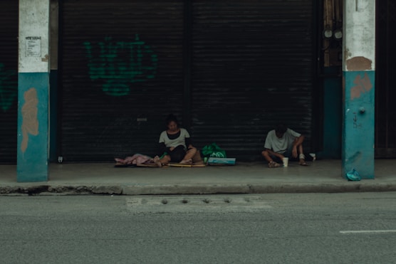 Two people sit on a sidewalk under a sheltered area in front of closed metal shutters. They appear to be surrounded by bags and blankets, suggesting they might be experiencing homelessness. The overall atmosphere is quiet and somber.