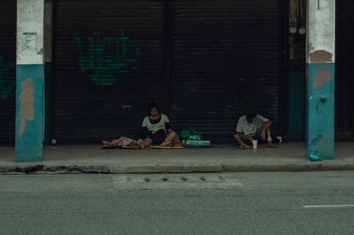 Two people sit on a sidewalk under a sheltered area in front of closed metal shutters. They appear to be surrounded by bags and blankets, suggesting they might be experiencing homelessness. The overall atmosphere is quiet and somber.