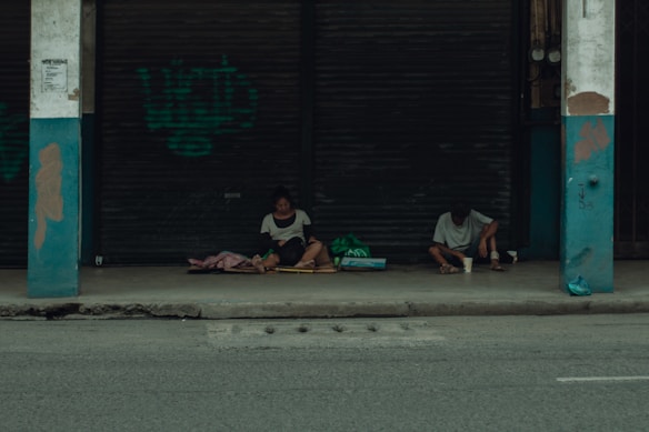 Two people sit on a sidewalk under a sheltered area in front of closed metal shutters. They appear to be surrounded by bags and blankets, suggesting they might be experiencing homelessness. The overall atmosphere is quiet and somber.