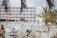 People are lounging by a poolside at a resort, with many colorful lounge chairs surrounding the area. The scene includes adults and children swimming and relaxing. A multi-story hotel building serves as the backdrop, and palm trees are present, giving a tropical ambiance.