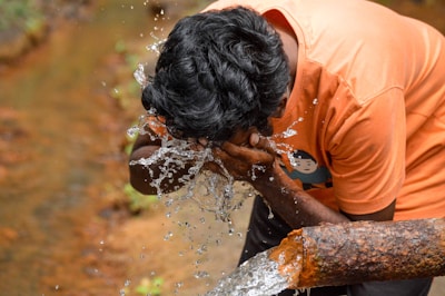 A person with dark hair is washing their face with clear water flowing from a rusted metal pipe. The individual is wearing an orange shirt, and the water is splashing visibly as it contacts their face. The background is a blur of earth tones, possibly indicating an outdoor, natural setting.