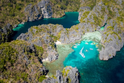 An aerial view of a beautiful lagoon surrounded by rugged limestone cliffs covered in greenery. Turquoise and deep blue waters fill the lagoon, with numerous small boats and kayaks scattered across the water. The water's clarity reveals shallow reefs and patches of sand.