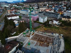 An aerial view of a construction site with scaffolding and machinery surrounded by a residential and commercial area. Buildings with varied architectural styles and rooftops spread across the landscape, set against a backdrop of distant snow-capped mountains. A prominent green construction crane stands amidst the site indicating ongoing development.