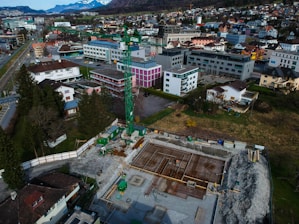 An aerial view of a construction site with scaffolding and machinery surrounded by a residential and commercial area. Buildings with varied architectural styles and rooftops spread across the landscape, set against a backdrop of distant snow-capped mountains. A prominent green construction crane stands amidst the site indicating ongoing development.