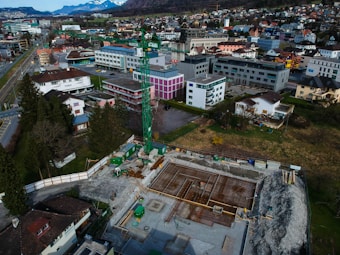 An aerial view of a construction site with scaffolding and machinery surrounded by a residential and commercial area. Buildings with varied architectural styles and rooftops spread across the landscape, set against a backdrop of distant snow-capped mountains. A prominent green construction crane stands amidst the site indicating ongoing development.