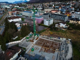 An aerial view of a construction site with scaffolding and machinery surrounded by a residential and commercial area. Buildings with varied architectural styles and rooftops spread across the landscape, set against a backdrop of distant snow-capped mountains. A prominent green construction crane stands amidst the site indicating ongoing development.