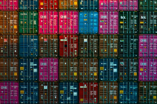 A dense arrangement of colorful shipping containers is stacked in a precise grid pattern. Containers in shades of blue, pink, red, green, and brown create a vivid mosaic, with various labels and logos visible on each.