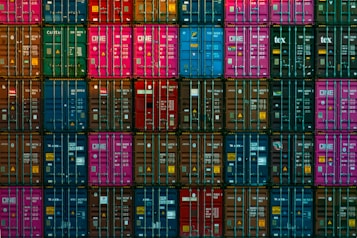A dense arrangement of colorful shipping containers is stacked in a precise grid pattern. Containers in shades of blue, pink, red, green, and brown create a vivid mosaic, with various labels and logos visible on each.