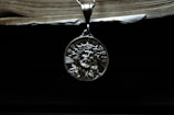 A silver pendant depicting a religious figure with a crown of thorns is hanging against a dark background. Above it, the edge of an old book with visible pages can be seen, casting a shadow.