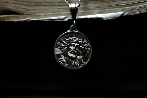 A silver pendant depicting a religious figure with a crown of thorns is hanging against a dark background. Above it, the edge of an old book with visible pages can be seen, casting a shadow.