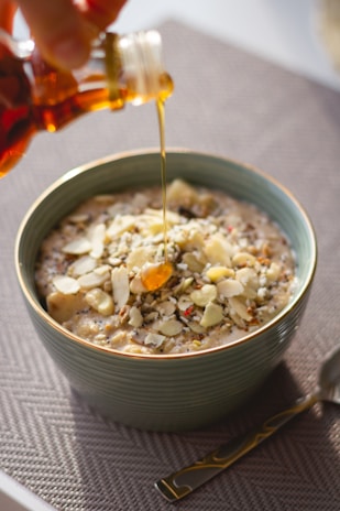 A bowl of oatmeal topped with sliced almonds and seeds is being drizzled with honey from a small bottle. The bowl is placed on a textured gray surface, and there is a gold-handled spoon next to it.