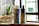 A bottle of CBD Pain Freeze spray and a dropper bottle of CBD Vanilla oil are placed on a wooden surface. Next to them is a small glass planter with a succulent, filled with pebbles and sand. The background features a softly blurred window, suggesting natural light entering the room.