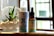 A bottle of CBD Pain Freeze spray and a dropper bottle of CBD Vanilla oil are placed on a wooden surface. Next to them is a small glass planter with a succulent, filled with pebbles and sand. The background features a softly blurred window, suggesting natural light entering the room.