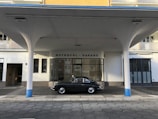 An elegant vintage car is parked in front of a retro-style building labeled 'Metropol-Garage'. The architecture features round columns painted with a touch of blue, with a canopy extending over the vehicle. The building has a mix of glass and solid walls with neutral colors.