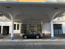 An elegant vintage car is parked in front of a retro-style building labeled 'Metropol-Garage'. The architecture features round columns painted with a touch of blue, with a canopy extending over the vehicle. The building has a mix of glass and solid walls with neutral colors.