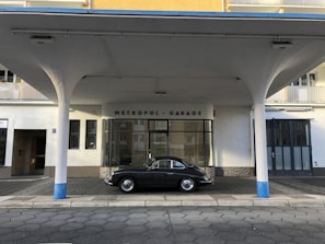 An elegant vintage car is parked in front of a retro-style building labeled 'Metropol-Garage'. The architecture features round columns painted with a touch of blue, with a canopy extending over the vehicle. The building has a mix of glass and solid walls with neutral colors.