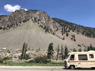 A rocky mountain slope covered with sparse pine trees under a clear blue sky. A beige and white recreational vehicle is parked on a roadside near the mountain. A few white clouds are visible in the sky.