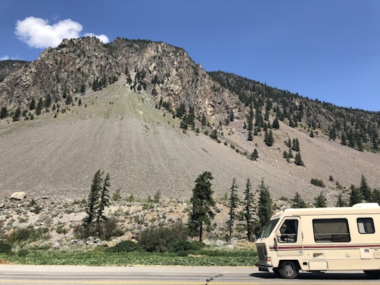 A rocky mountain slope covered with sparse pine trees under a clear blue sky. A beige and white recreational vehicle is parked on a roadside near the mountain. A few white clouds are visible in the sky.
