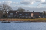 A picturesque lakeside house with a rustic charm, surrounded by bare trees and a grassy landscape. The building features a red-tiled roof and multiple windows, typical of a countryside dwelling. The weather is clear with some clouds in the sky, suggesting a calm and peaceful environment.