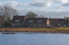 A picturesque lakeside house with a rustic charm, surrounded by bare trees and a grassy landscape. The building features a red-tiled roof and multiple windows, typical of a countryside dwelling. The weather is clear with some clouds in the sky, suggesting a calm and peaceful environment.