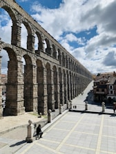 A stone aqueduct with multiple arches stretches across a cityscape under a partly cloudy sky. The ancient structure towers over a paved open area, where a few people are visible walking or resting. The scene captures a blend of historical architecture and urban life, with buildings visible in the background.