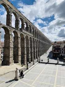 A stone aqueduct with multiple arches stretches across a cityscape under a partly cloudy sky. The ancient structure towers over a paved open area, where a few people are visible walking or resting. The scene captures a blend of historical architecture and urban life, with buildings visible in the background.
