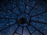 A person is seated with their legs forward, positioned at the center of a circular structure that resembles a spacecraft cockpit. Surrounding the structure are streaks of blue and white light, creating a sense of high-speed motion through space.