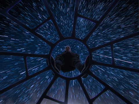 A person is seated with their legs forward, positioned at the center of a circular structure that resembles a spacecraft cockpit. Surrounding the structure are streaks of blue and white light, creating a sense of high-speed motion through space.