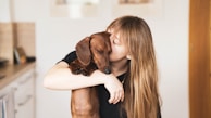 A person with long hair is affectionately holding and kissing a brown dachshund dog. The background is a softly lit indoor setting with blurred kitchen elements and a wooden door visible.