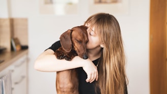 A person with long hair is affectionately holding and kissing a brown dachshund dog. The background is a softly lit indoor setting with blurred kitchen elements and a wooden door visible.