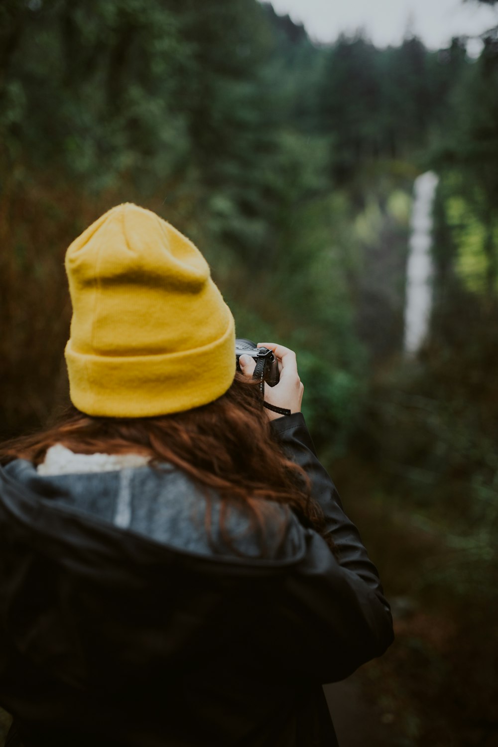 pessoa em boné de malha amarela e jaqueta preta tirando foto de árvores verdes durante o dia