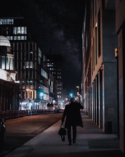 A lone figure walking along a city roadside under a starry night sky, deep in thought.