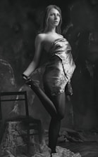 A black and white portrait of a young woman posed gracefully with one leg raised, standing next to a wooden chair. She is draped in a patterned cloth that wraps around her upper body and flows down. The background is dark and textured, adding depth to the scene.