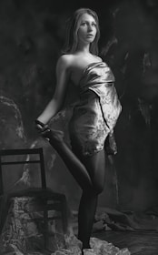 A black and white portrait of a young woman posed gracefully with one leg raised, standing next to a wooden chair. She is draped in a patterned cloth that wraps around her upper body and flows down. The background is dark and textured, adding depth to the scene.