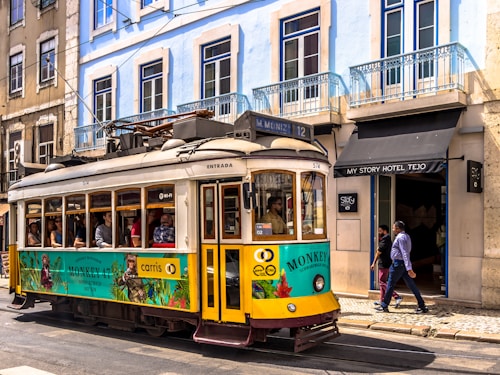 A vintage yellow tram travels down a street lined with historic buildings. People can be seen inside the tram, indicating it is in operation. The building to the right features a hotel entrance with people entering and exiting. The exterior of the buildings is characterized by pastel colors and wrought iron balconies, typical of European architecture.