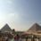 A large number of people gather near ancient Egyptian landmarks under a clear blue sky. Two pyramids stand prominently in the background with the Sphinx visible nearby.
