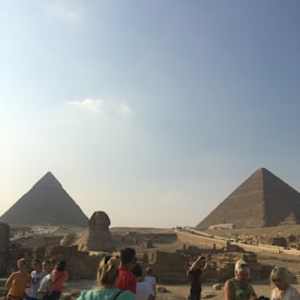 A large number of people gather near ancient Egyptian landmarks under a clear blue sky. Two pyramids stand prominently in the background with the Sphinx visible nearby.