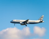 A commercial airplane with the branding of Aegean Airlines is in mid-air against a backdrop of a clear blue sky and a few fluffy white clouds. The aircraft is lit by natural daylight, showcasing its white fuselage and the distinctive blue accents.