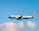 A commercial airplane with the branding of Aegean Airlines is in mid-air against a backdrop of a clear blue sky and a few fluffy white clouds. The aircraft is lit by natural daylight, showcasing its white fuselage and the distinctive blue accents.
