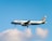 A commercial airplane with the branding of Aegean Airlines is in mid-air against a backdrop of a clear blue sky and a few fluffy white clouds. The aircraft is lit by natural daylight, showcasing its white fuselage and the distinctive blue accents.