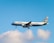 A commercial airplane with the branding of Aegean Airlines is in mid-air against a backdrop of a clear blue sky and a few fluffy white clouds. The aircraft is lit by natural daylight, showcasing its white fuselage and the distinctive blue accents.