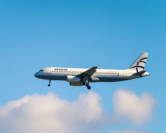 A commercial airplane with the branding of Aegean Airlines is in mid-air against a backdrop of a clear blue sky and a few fluffy white clouds. The aircraft is lit by natural daylight, showcasing its white fuselage and the distinctive blue accents.