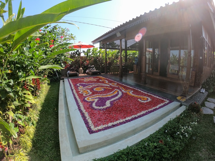 A lush garden setting featuring a flower petal arrangement in vibrant colors on the ground. The arrangement is bordered by neatly trimmed grass and surrounded by tropical plants. A wooden building with a porch and curtains is situated in the background, accompanied by an orange patio umbrella and lounge chairs.