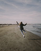 man in black jacket and blue denim jeans jumping on beach during daytime