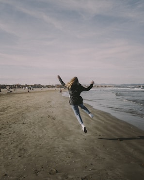 man in black jacket and blue denim jeans jumping on beach during daytime