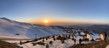 A breathtaking landscape of a snowy mountain at sunset. The sun is setting on the horizon, casting an orange glow across the sky and illuminating the snow-covered slopes. There are ski lifts visible on the left, with a few people and trees dotting the forefront. Wooden signs are placed along a path, and the distant hills create a serene backdrop.