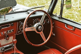 A vintage car interior featuring a classic wooden steering wheel and red leather upholstery. The dashboard includes analog dials and switches, with chrome accents that add to the retro aesthetic. The window handle and side mirror reflect the classic car design.