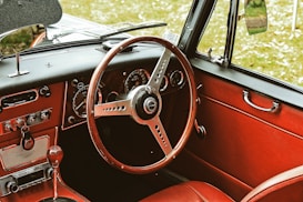 A vintage car interior featuring a classic wooden steering wheel and red leather upholstery. The dashboard includes analog dials and switches, with chrome accents that add to the retro aesthetic. The window handle and side mirror reflect the classic car design.