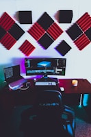 A modern gaming or editing setup featuring a wide monitor with vibrant displays, surrounded by acoustic foam panels on the wall in black and red. On the desk, there is a laptop, a keyboard, a mouse, and a candle adding ambiance. The setup includes colorful LED lighting under the desk and a cushioned chair labeled Karnox.