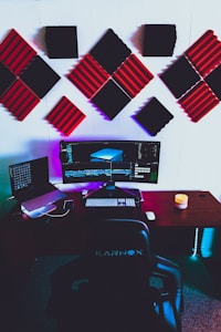 A modern gaming or editing setup featuring a wide monitor with vibrant displays, surrounded by acoustic foam panels on the wall in black and red. On the desk, there is a laptop, a keyboard, a mouse, and a candle adding ambiance. The setup includes colorful LED lighting under the desk and a cushioned chair labeled Karnox.